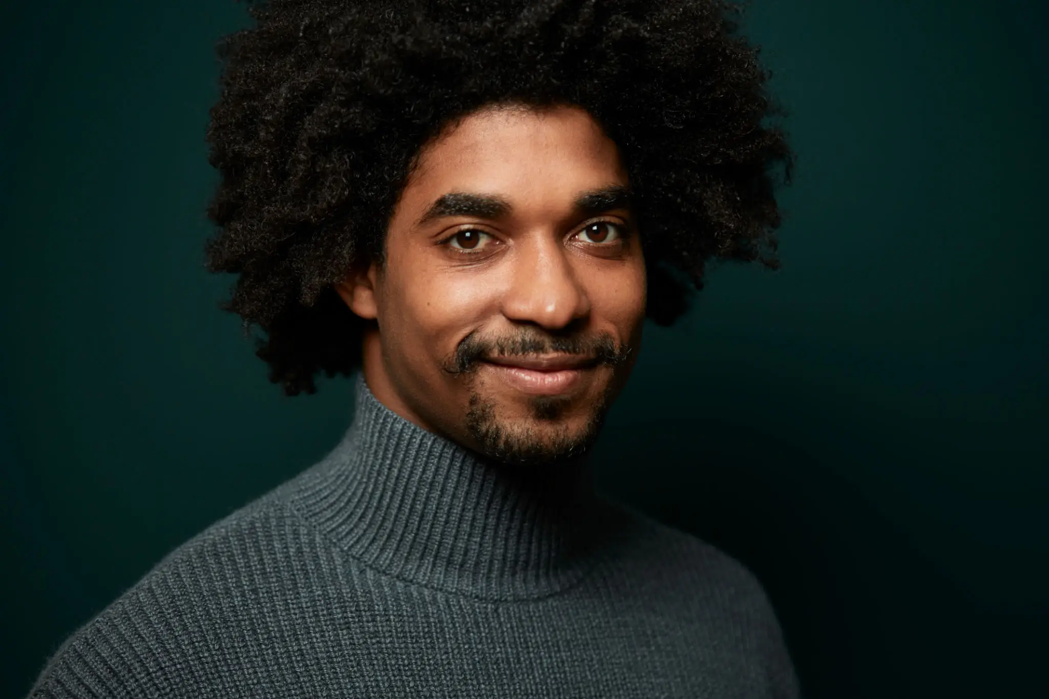 Man smiling in gray turtleneck against green background.